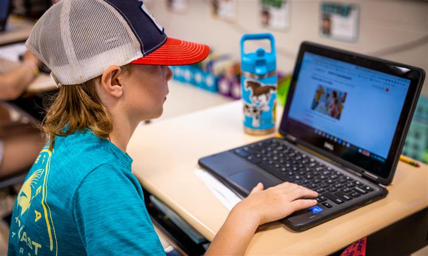 A boy wearing a red cap uses a Chromebook on his desk